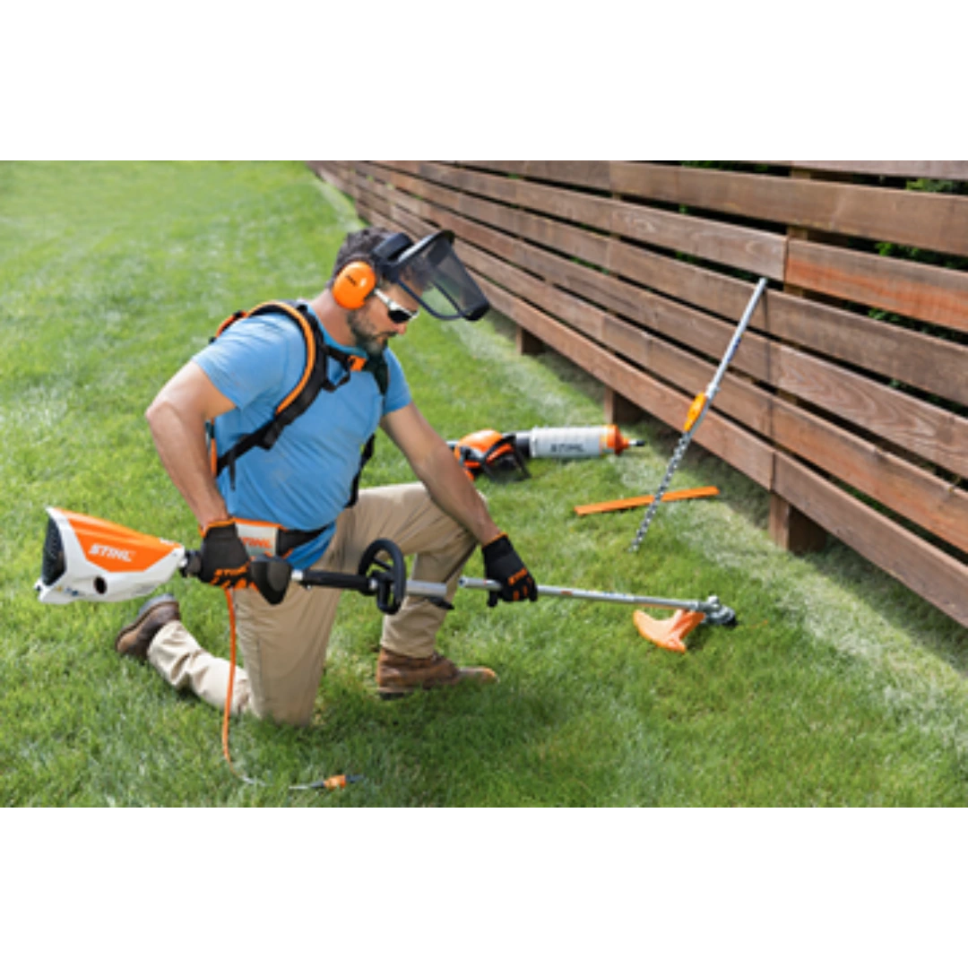 Man wearing protective gear using a battery-powered string trimmer on grass near wooden fence in outdoor power tools setting