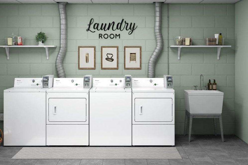 Row of white commercial electric dryers with coin slide controls in a light green laundry room with sink and wall shelves