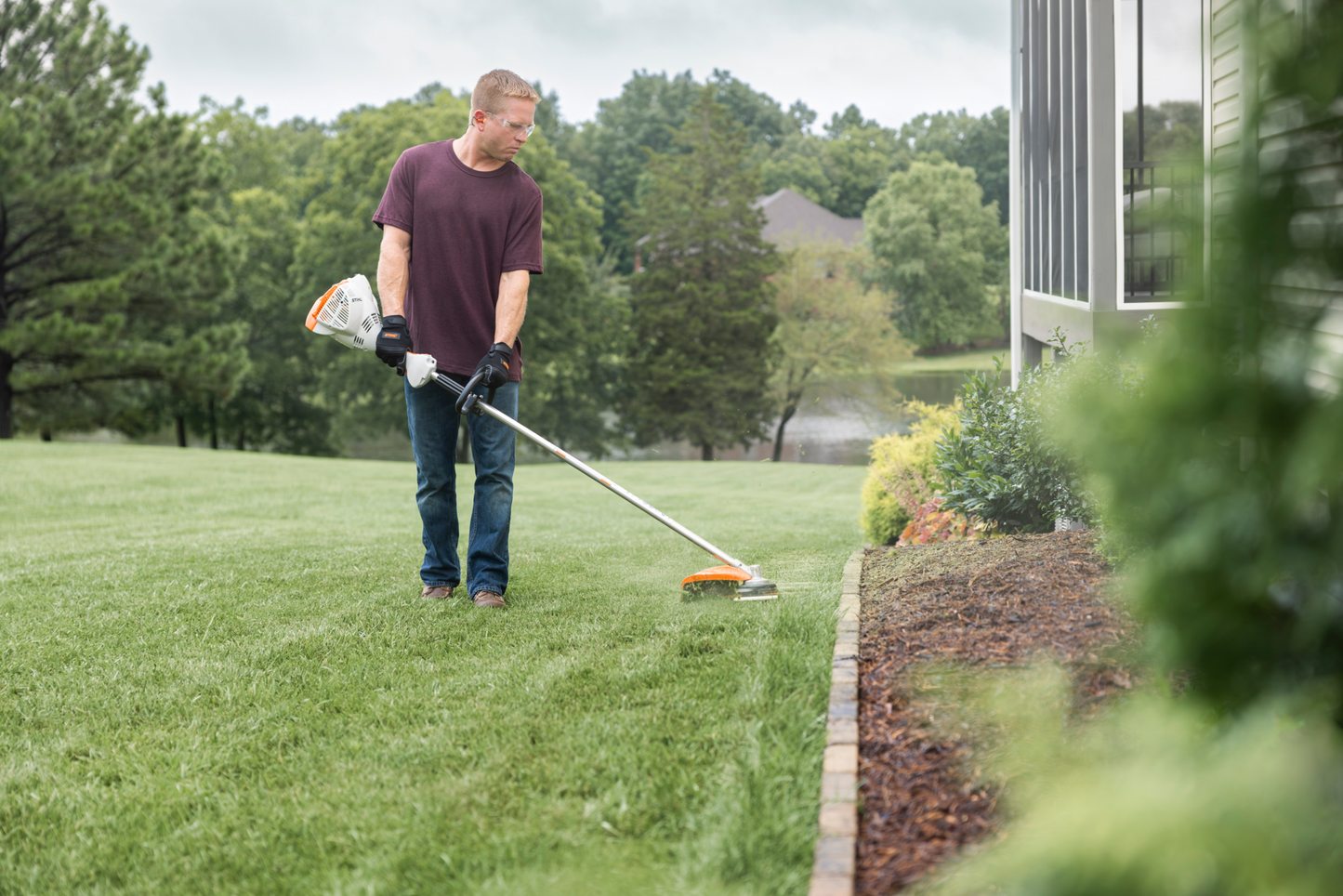 Person using a straight shaft loop-handle trimmer to edge a lawn near a landscaped garden for yard maintenance.