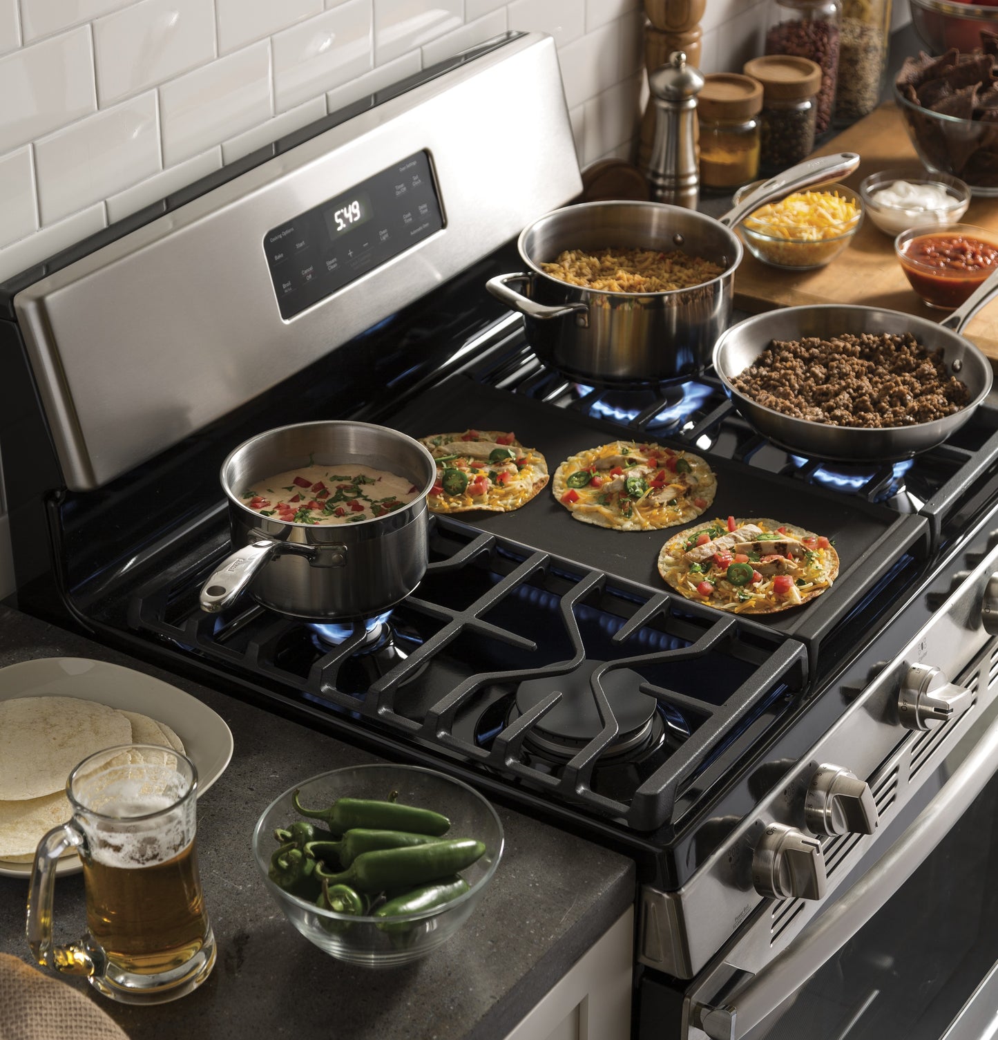 Stainless steel gas range with multiple burners cooking small pizzas, soup, ground beef, and rice in a kitchen setting