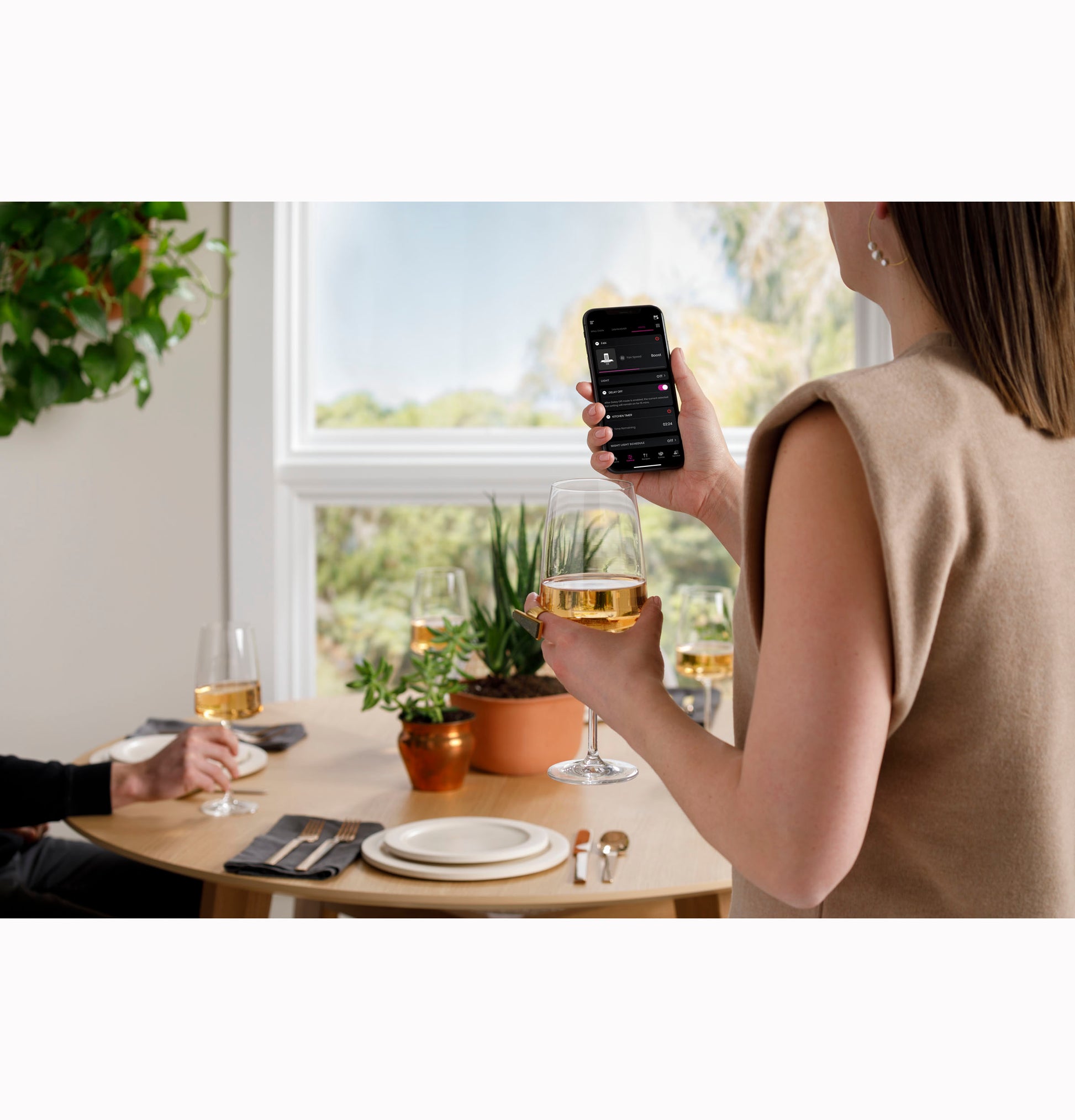 Person holding smartphone and wine glass by a round table set for dining with plants in a bright kitchen scene