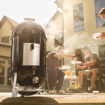 Black vertical charcoal smoker releasing smoke with people enjoying outdoor grilling and socializing in backyard setting