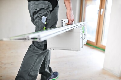 Person carrying a Festool guide rail and tool box in a bright room for woodworking and home improvement tasks