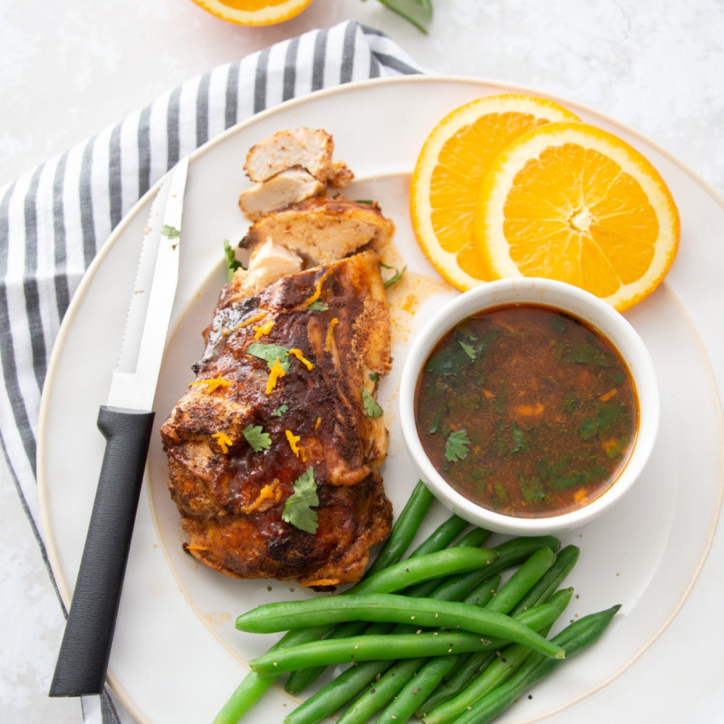 Serrated steak knife with black handle next to seasoned cooked chicken, green beans, dipping sauce, and orange slices on plate