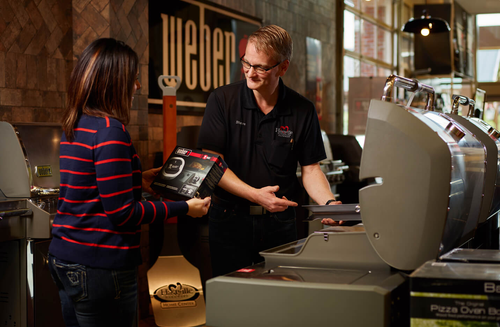 A store employee wearing a black shirt explains features of a stainless steel grill to a customer in a showroom with warm lighting.