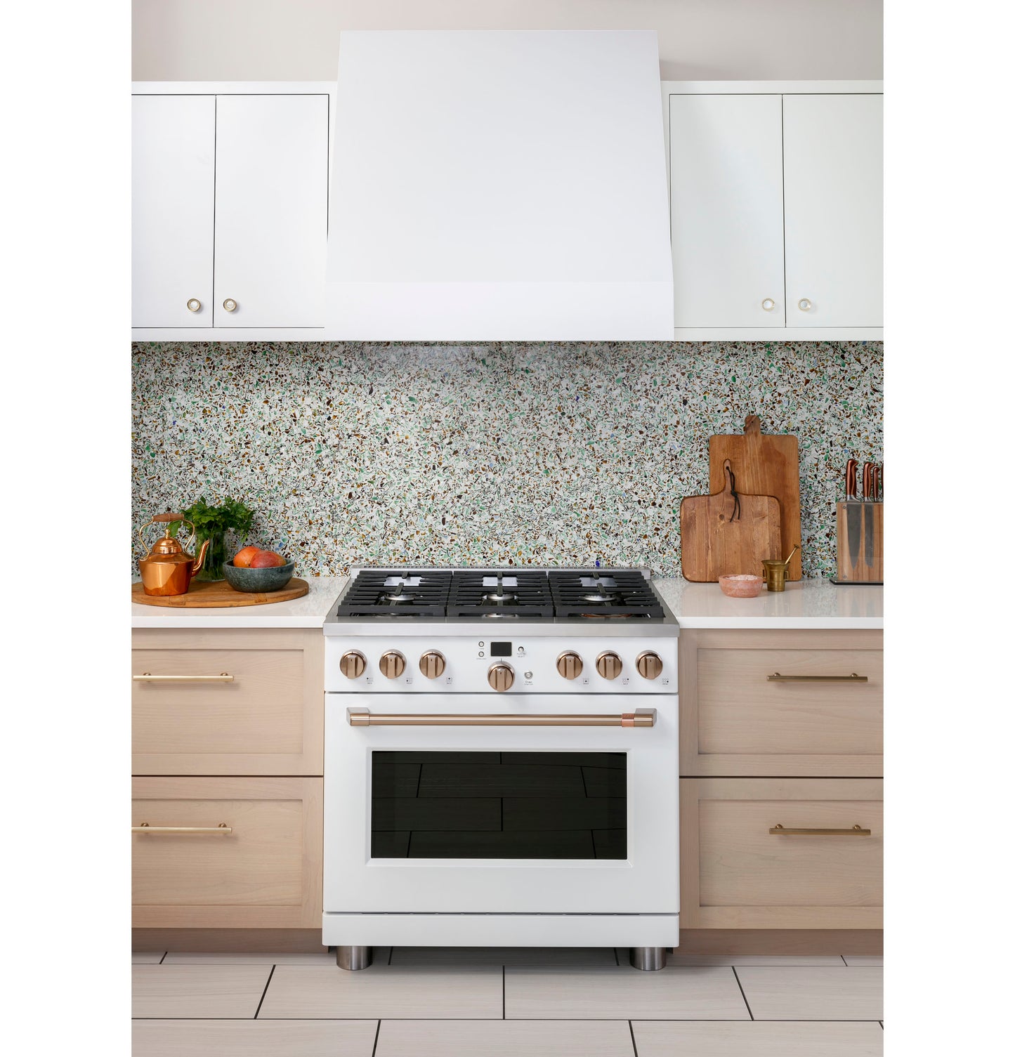 White gas range with six burners and brass knobs set between light wood drawers under white cabinetry and speckled backsplash