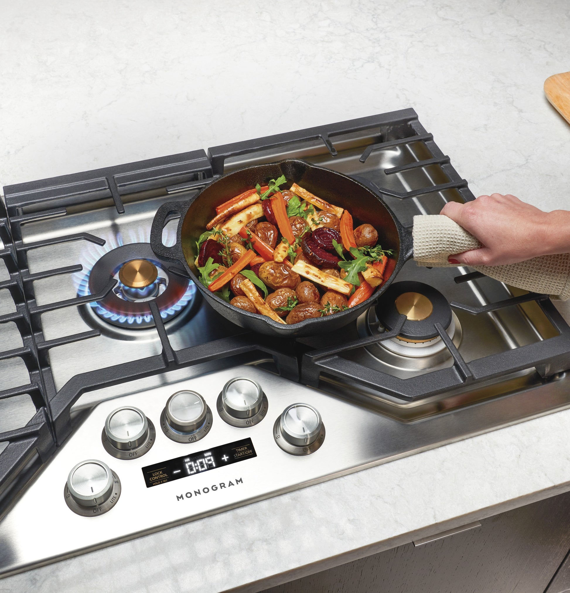 Cast iron skillet cooking vegetables on stainless steel deep-recessed gas cooktop with brass burners and control knobs