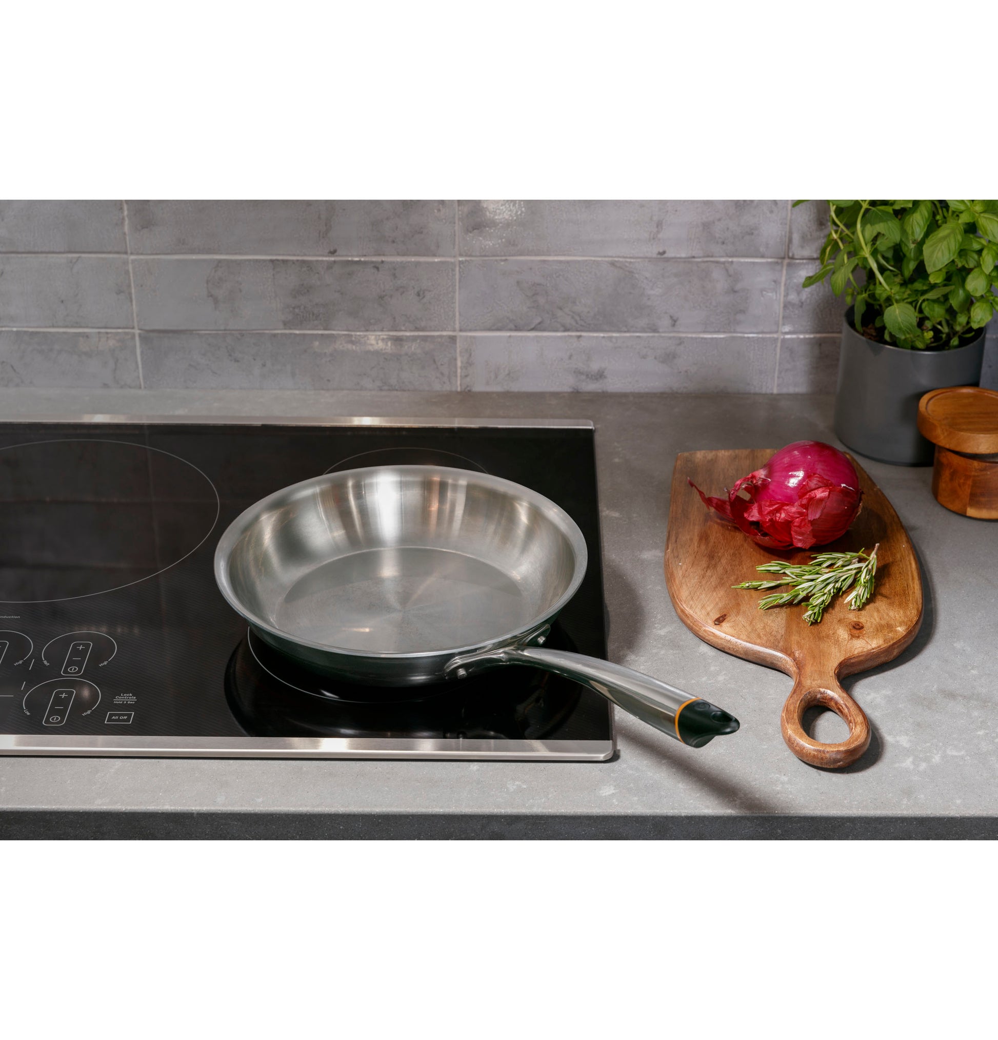 Stainless steel frying pan heating on black glass induction cooktop next to wooden cutting board with onion and herbs