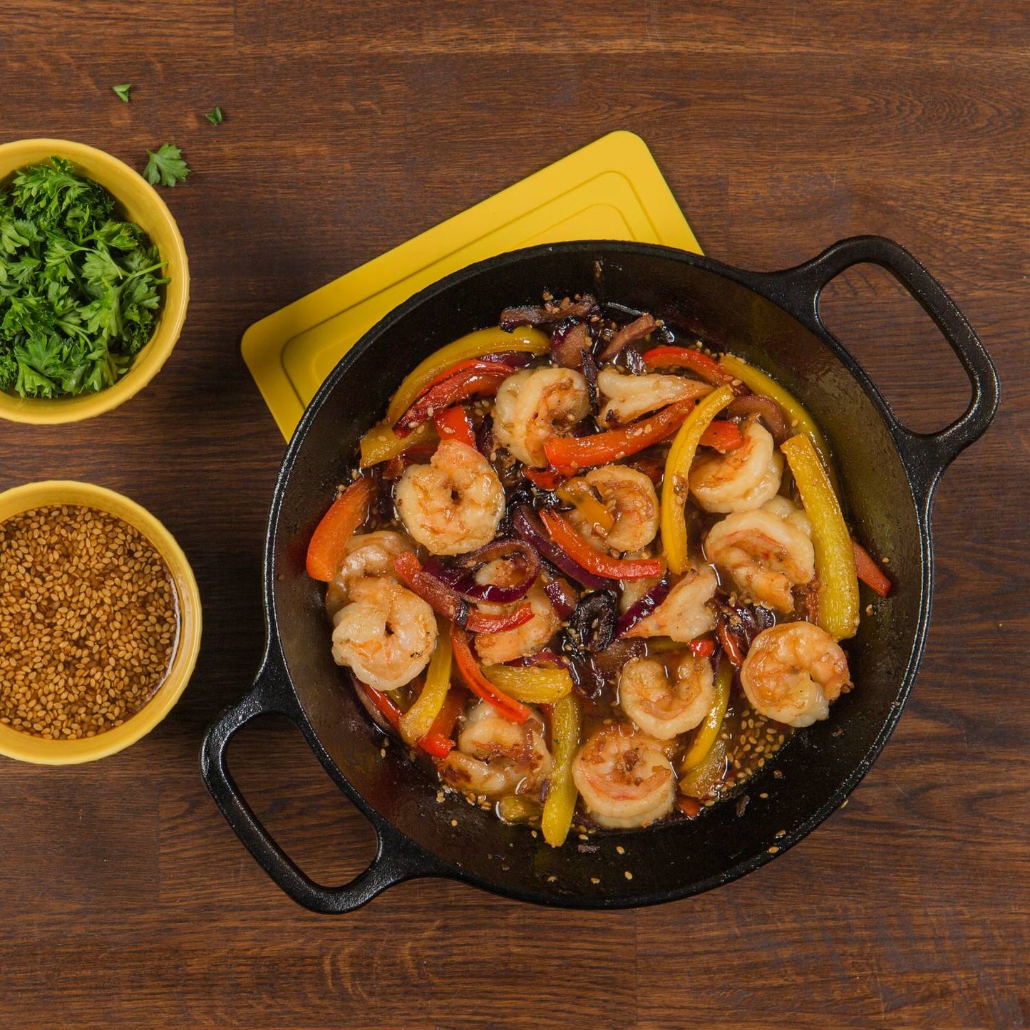 Round cast iron pan with shrimp and colorful bell peppers on wooden table beside bowls of herbs and sesame seeds in kitchenware collection
