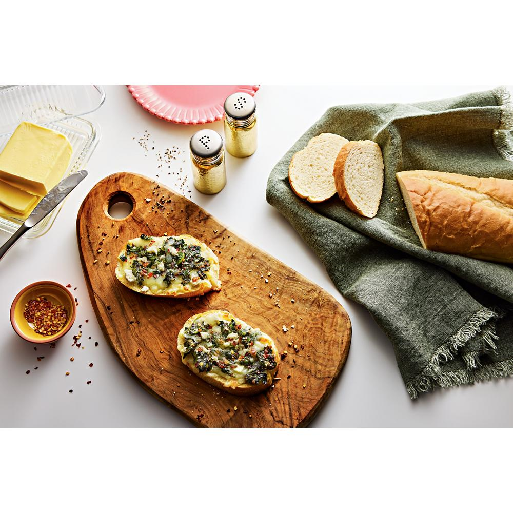 Two pieces of prepared bread with toppings on a wooden cutting board beside sliced loaf and seasonings