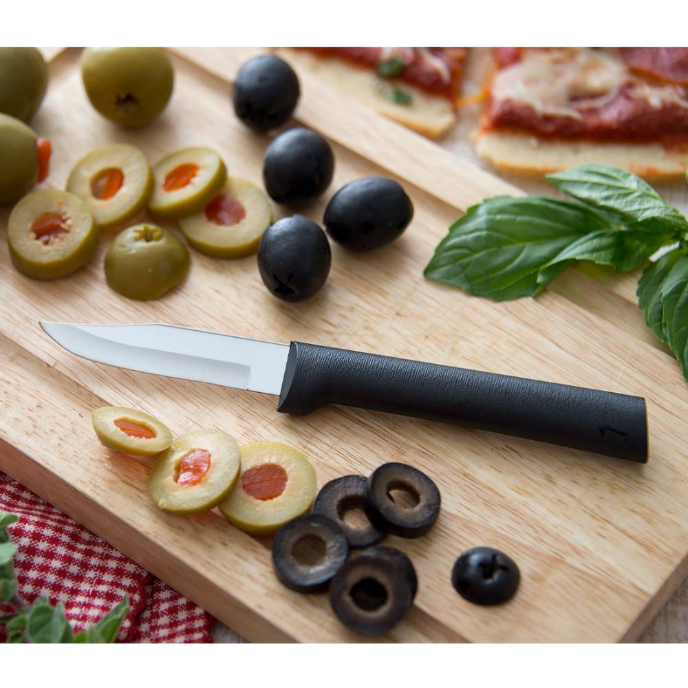 Sharp paring knife with black handle resting on a wooden board surrounded by sliced green and black olives and fresh basil leaves