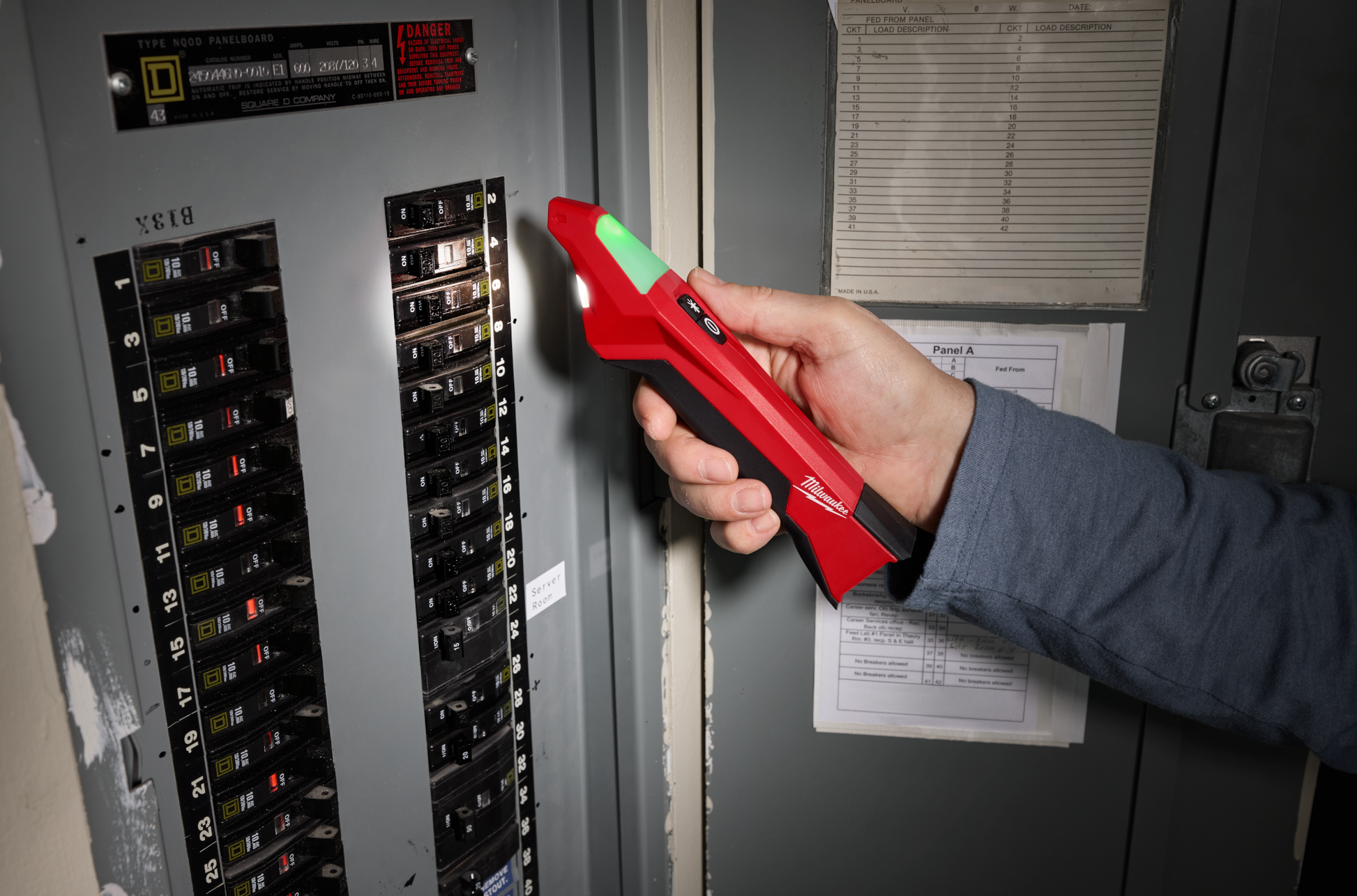 Person using red Milwaukee circuit breaker finder with LED light near electrical panel for home improvement tasks