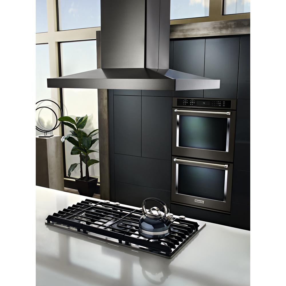 Stainless steel island canopy hood with built-in controls above gas cooktop in modern kitchen with double ovens