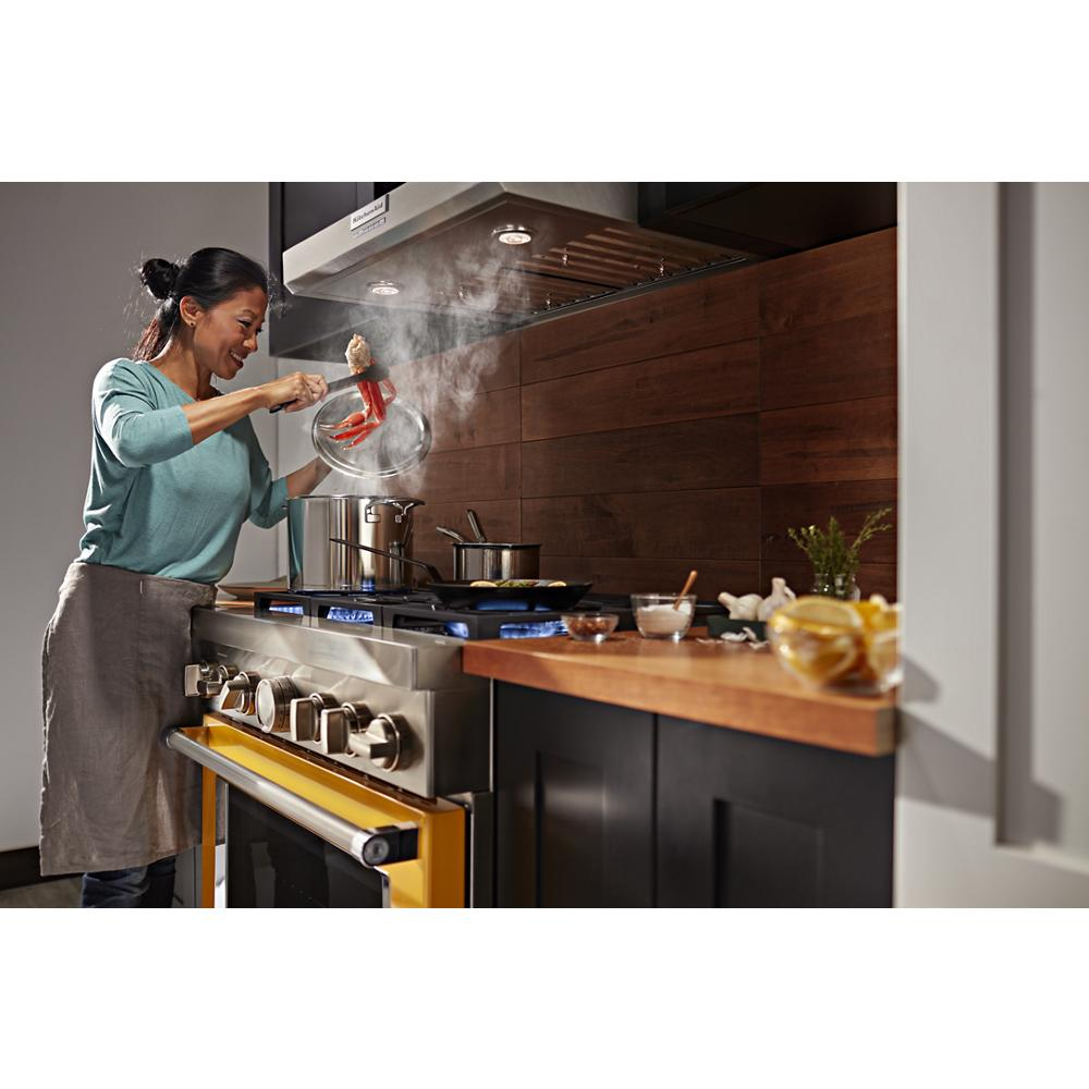 Woman cooking on yellow stove under stainless steel commercial-style wall-mount canopy range hood in modern kitchen