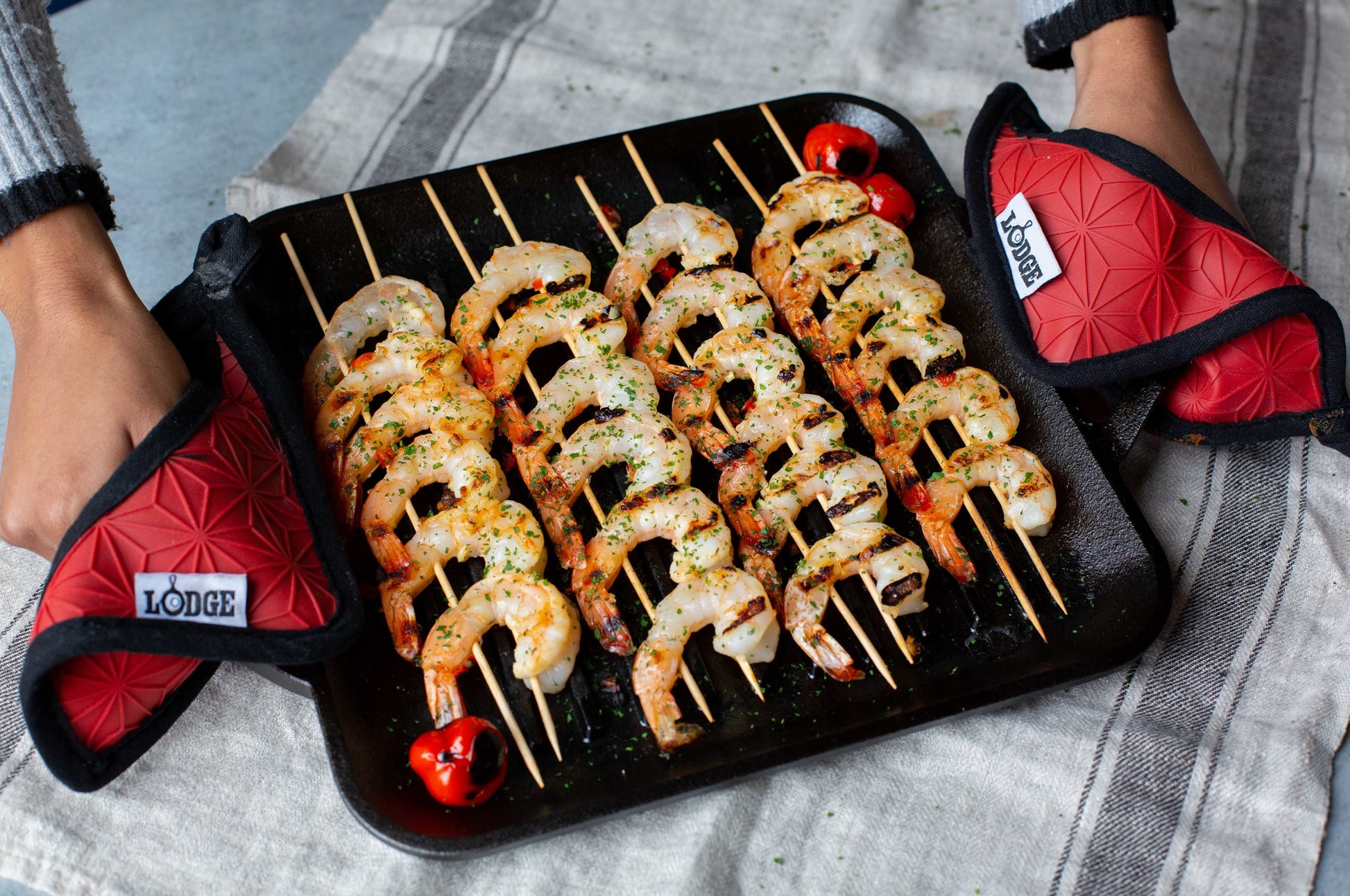 Black square cast iron grill pan with shrimp skewers and red Lodge branded heat-resistant mitts on fabric surface
