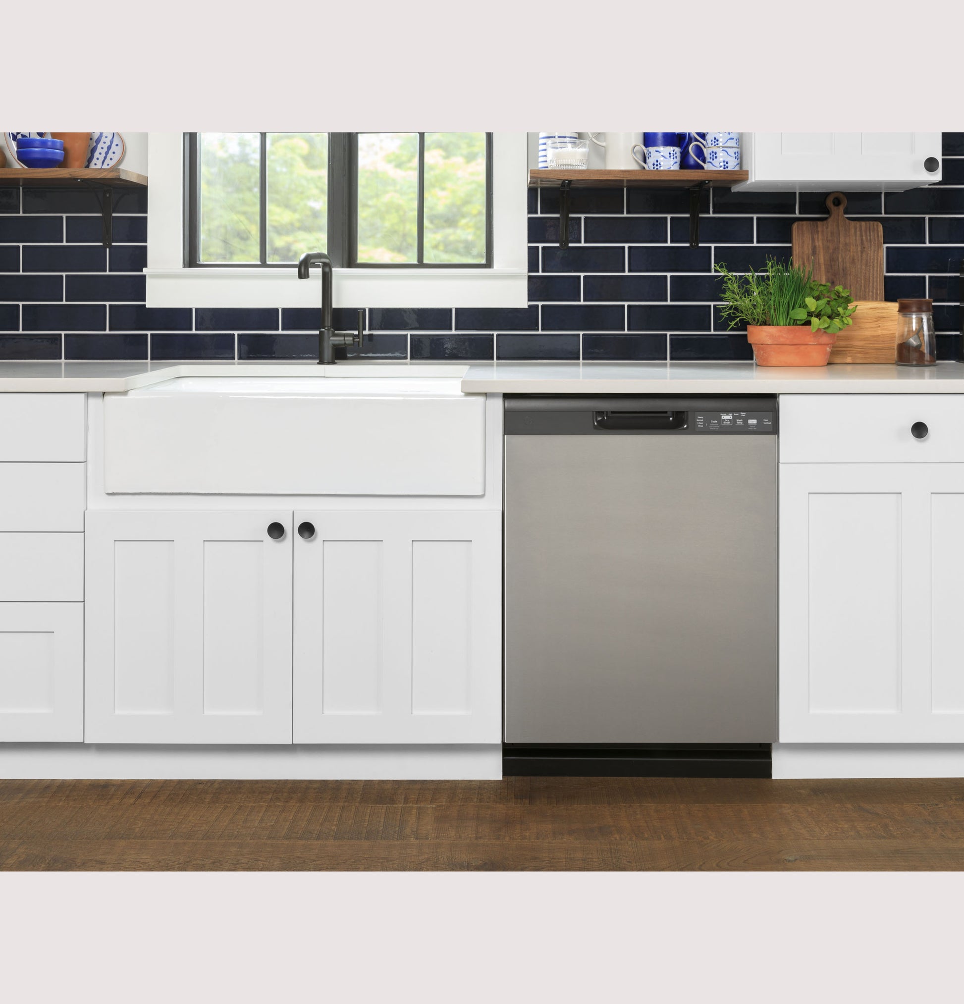 Stainless steel dishwasher with front controls integrated in modern white kitchen with farmhouse sink and dark tile backsplash
