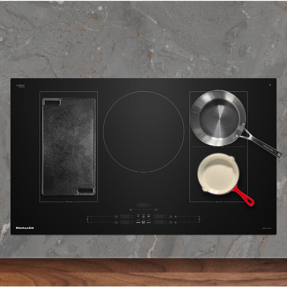 Black induction cooktop with five cooking zones including griddle, stainless steel pan, and cream ceramic skillet on granite counter