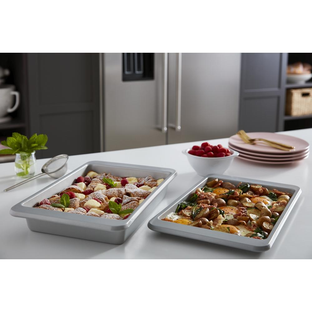 Two baking trays with pastries and roasted potatoes on white kitchen countertop near plates and raspberry bowl