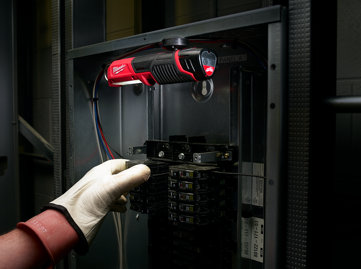Gloved hand working on electrical panel illuminated by compact red and black stick light from Milwaukee tools collection