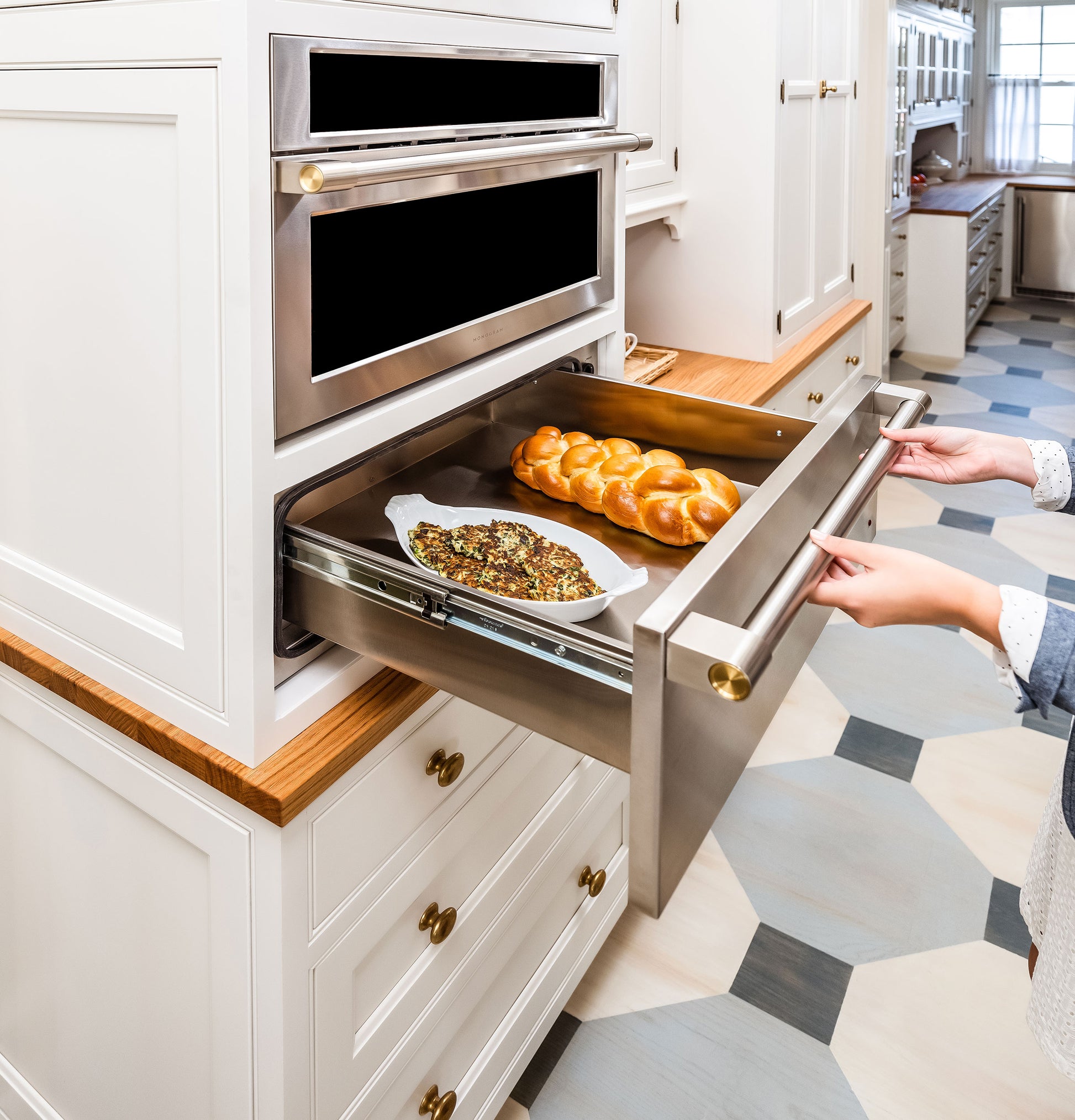Hands opening stainless steel warming drawer integrated into white kitchen cabinetry with baked goods inside