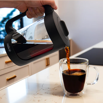 Hand pouring freshly brewed coffee from black-handled glass immersion coffee maker into clear double-walled cup on kitchen counter