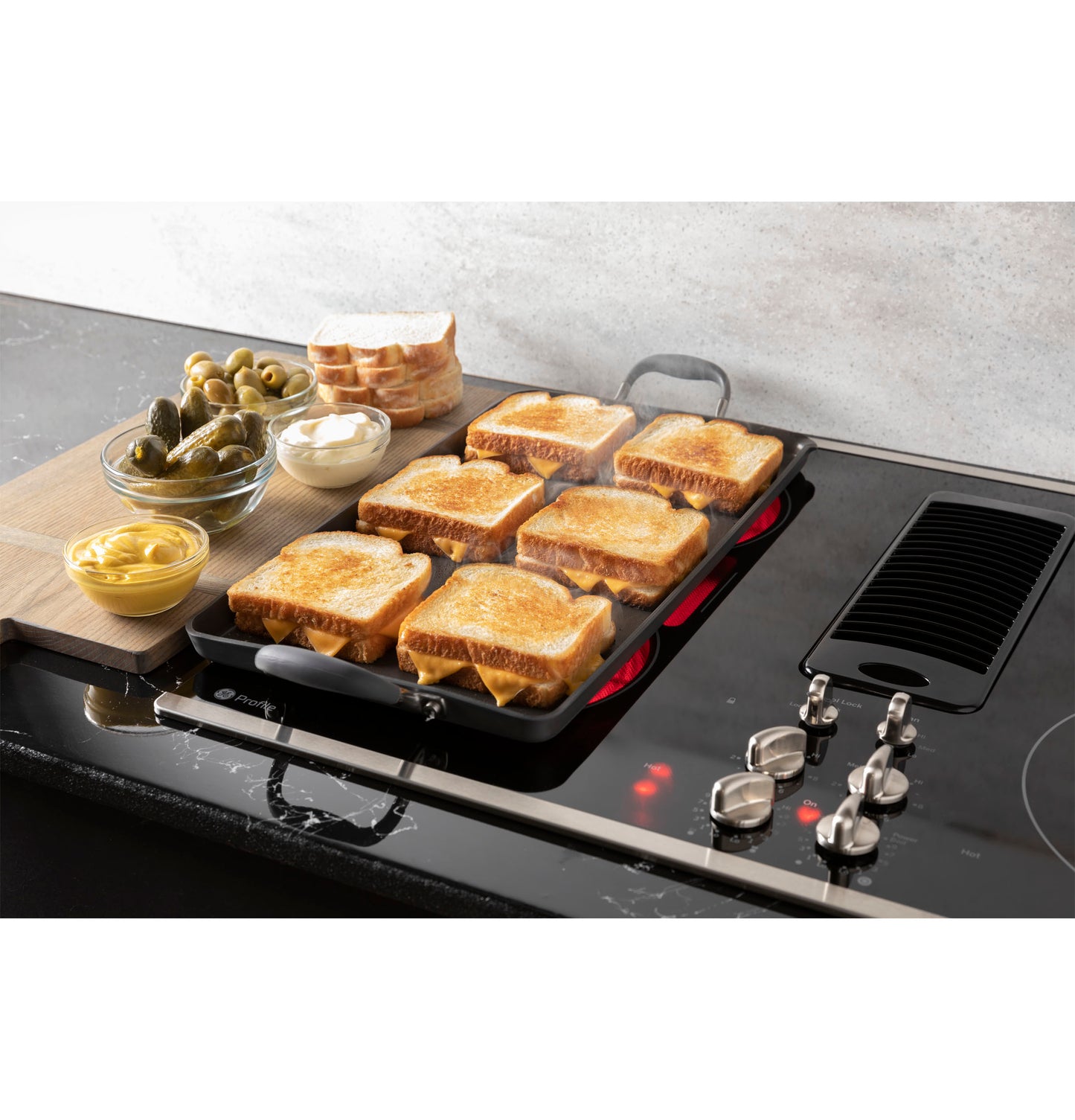 GE Profile downdraft electric cooktop with six toasted sandwiches on grill and condiments nearby in kitchenware setting