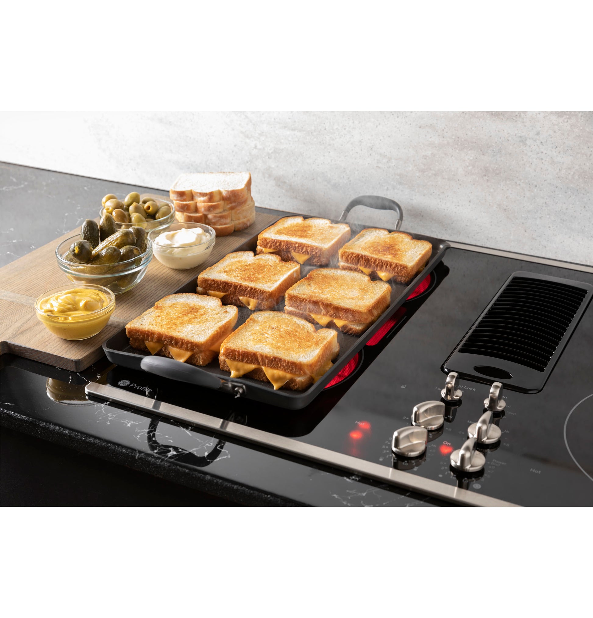 GE Profile downdraft electric cooktop with six toasted sandwiches on grill and condiments nearby in kitchenware setting