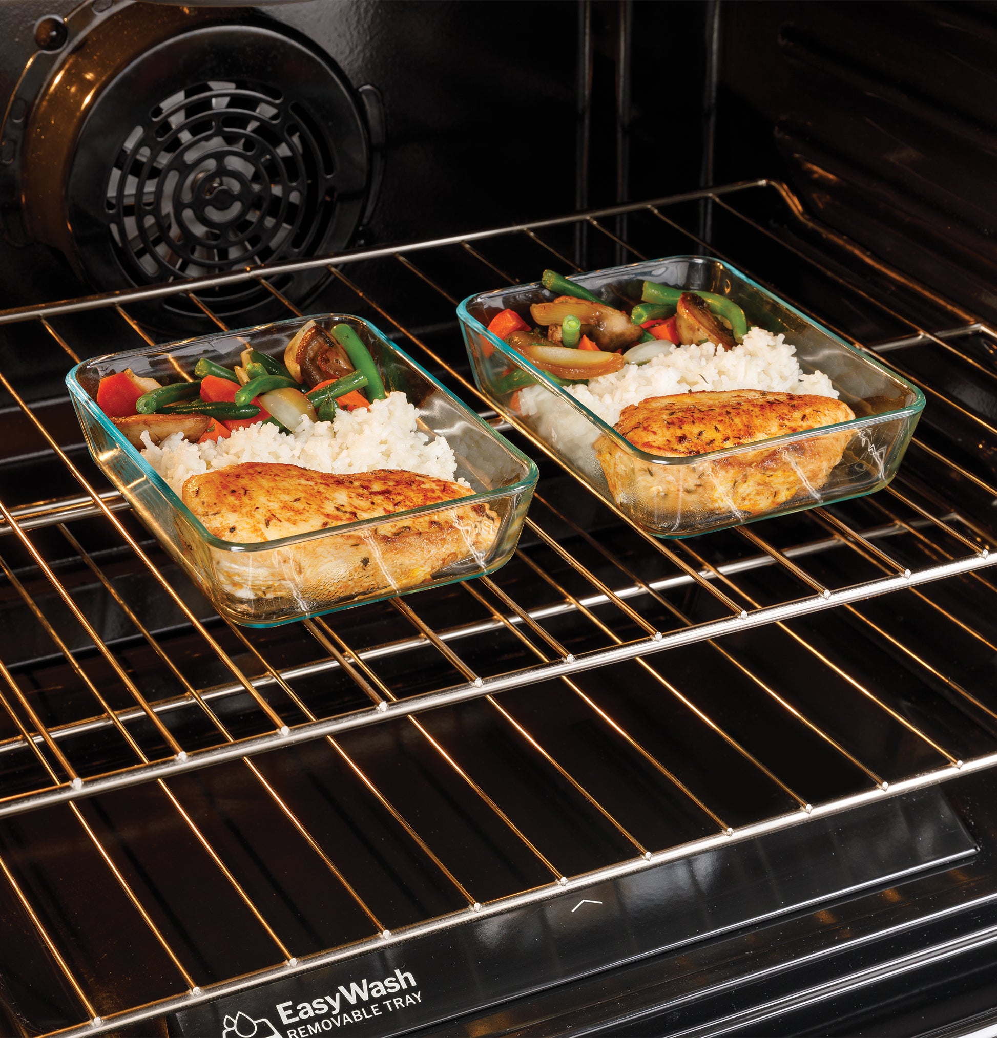 Two glass meal prep containers with chicken, rice, and vegetables inside convection oven with EasyWash tray beneath rack