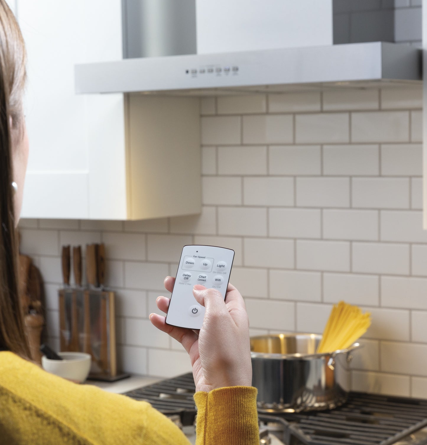 Person using remote control to adjust settings on a wall mount range hood above stovetop with pot and pasta inside