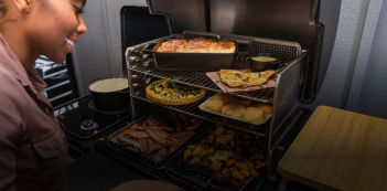 Person checking multiple shelves of cooked food inside a large pellet grill in outdoor living and grilling collections