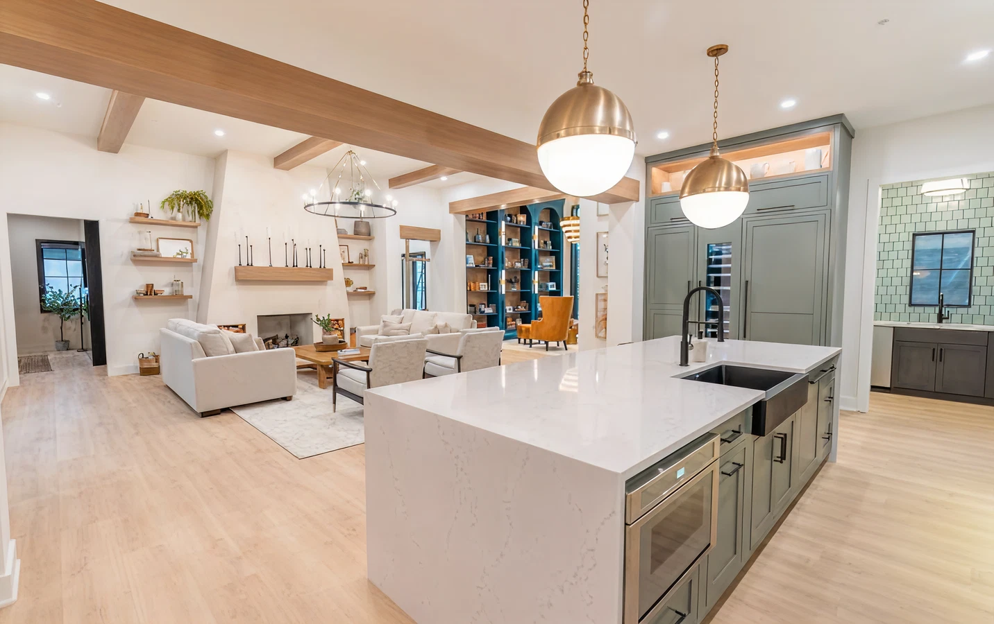 Open-concept living area with a modern kitchen island featuring pendant lights, a white countertop, sage green cabinetry, and seating adjacent to a cozy sitting room with neutral furniture and built-in shelves.