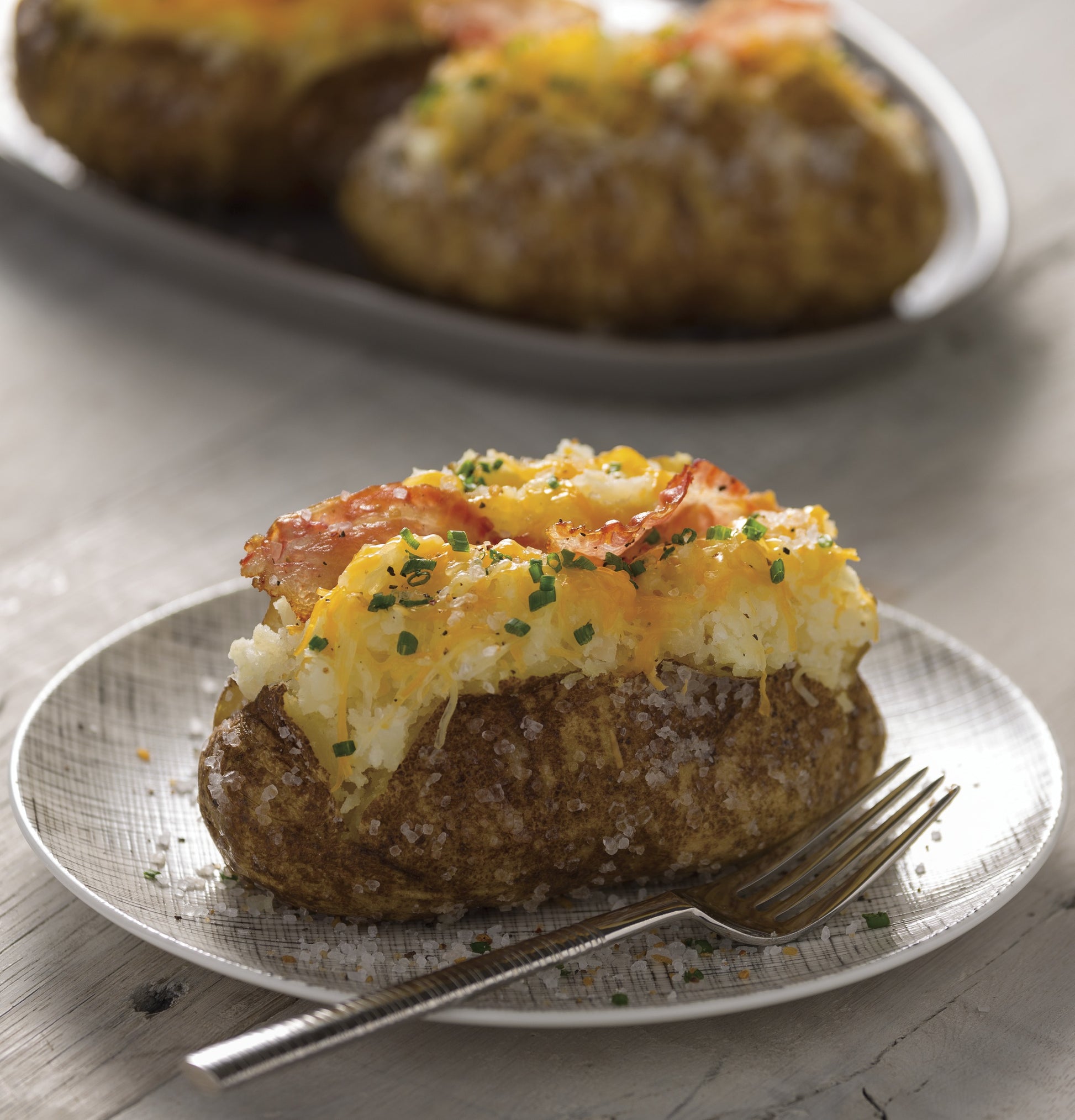 Baked stuffed potato topped with melted cheese, bacon, and chopped chives on a white plate with fork nearby