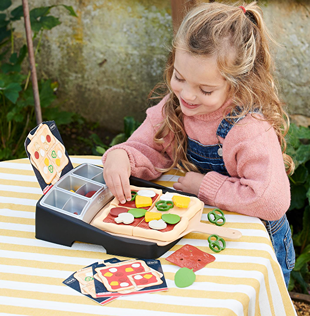 Child playing with Casdon Ooni pizza topping station toy featuring plastic toppings and recipe cards outdoors