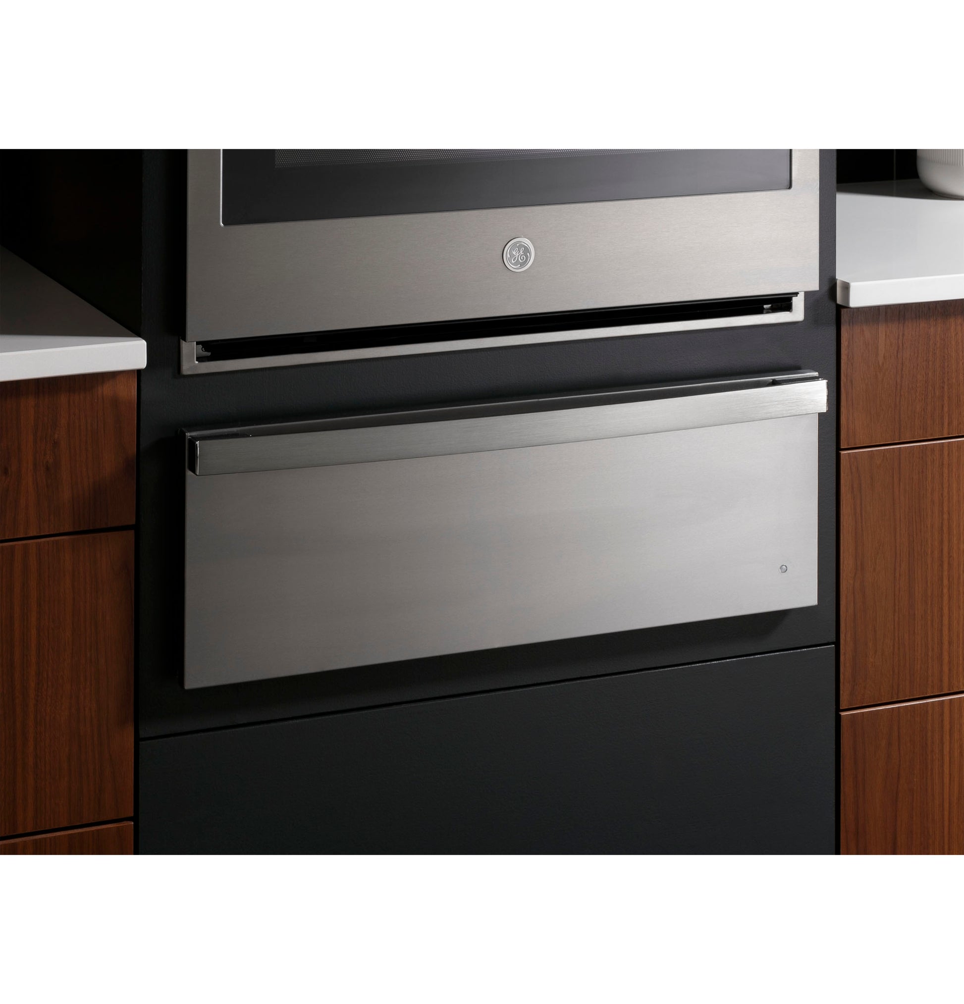 Built-in stainless steel warming drawer beneath a wall oven in kitchen cabinetry with wood finishes and black trim