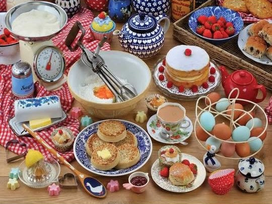 Vintage baking setup with measuring scale, mixing bowl, hand mixer, English muffins, eggs, and assorted pastries on red checkered cloth