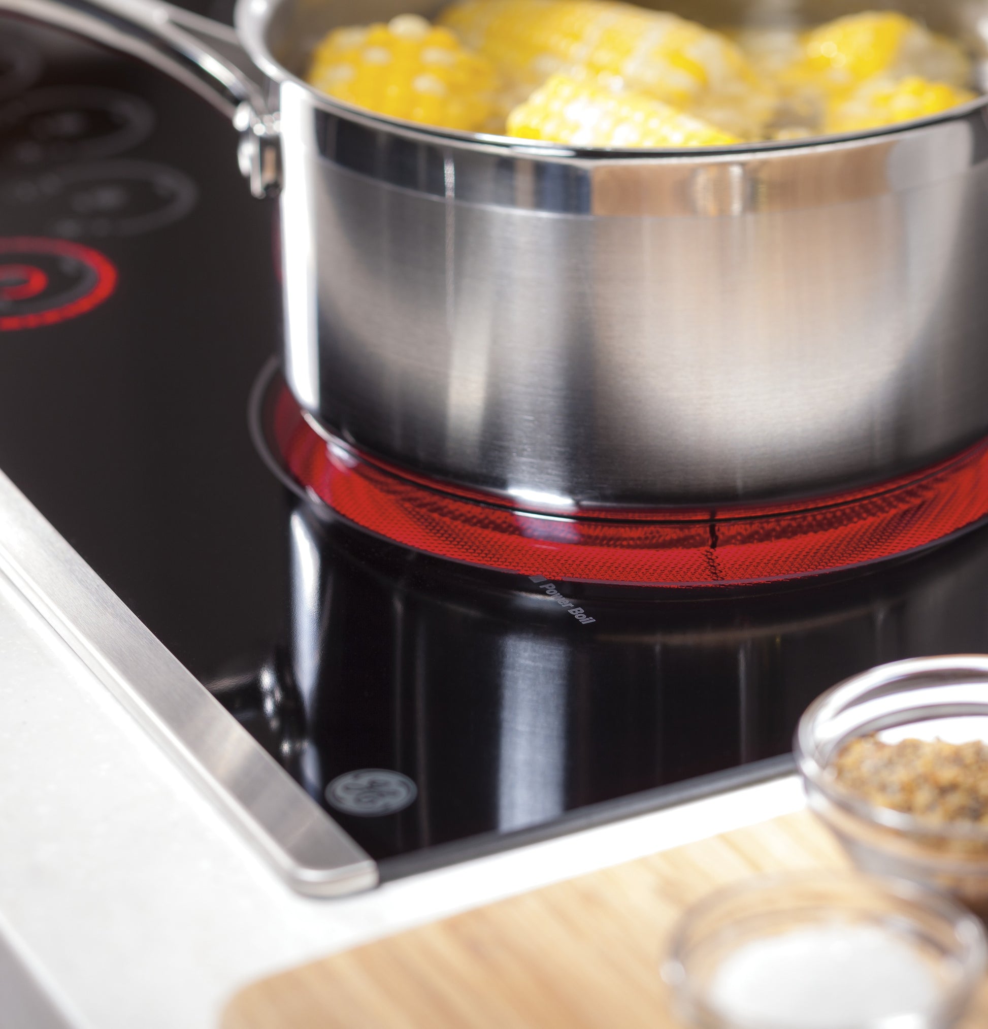 Close-up of stainless steel pot with corn boiling on active red heating element of a black glass electric cooktop in kitchenware collection