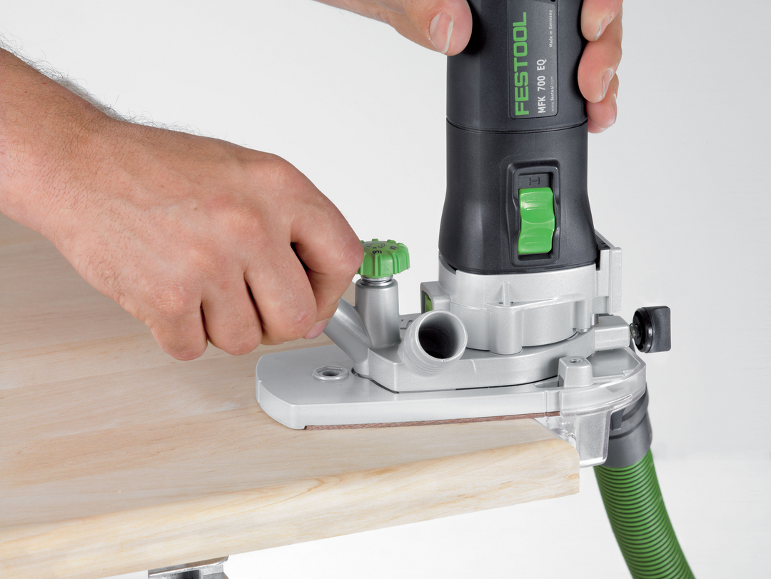 Person tightening green adjustment knob on trim router with attached dust extraction hose for woodworking tasks