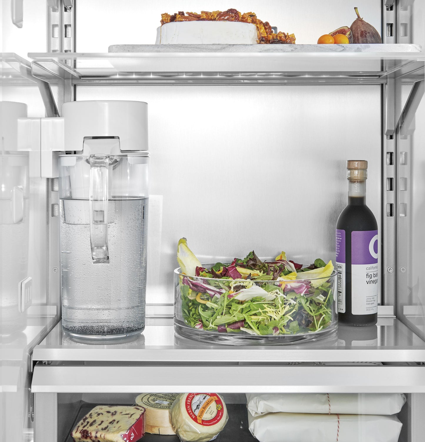 Interior view of panel-ready refrigerator with clear water pitcher, fresh salad, and bottled condiment in kitchen appliances collection