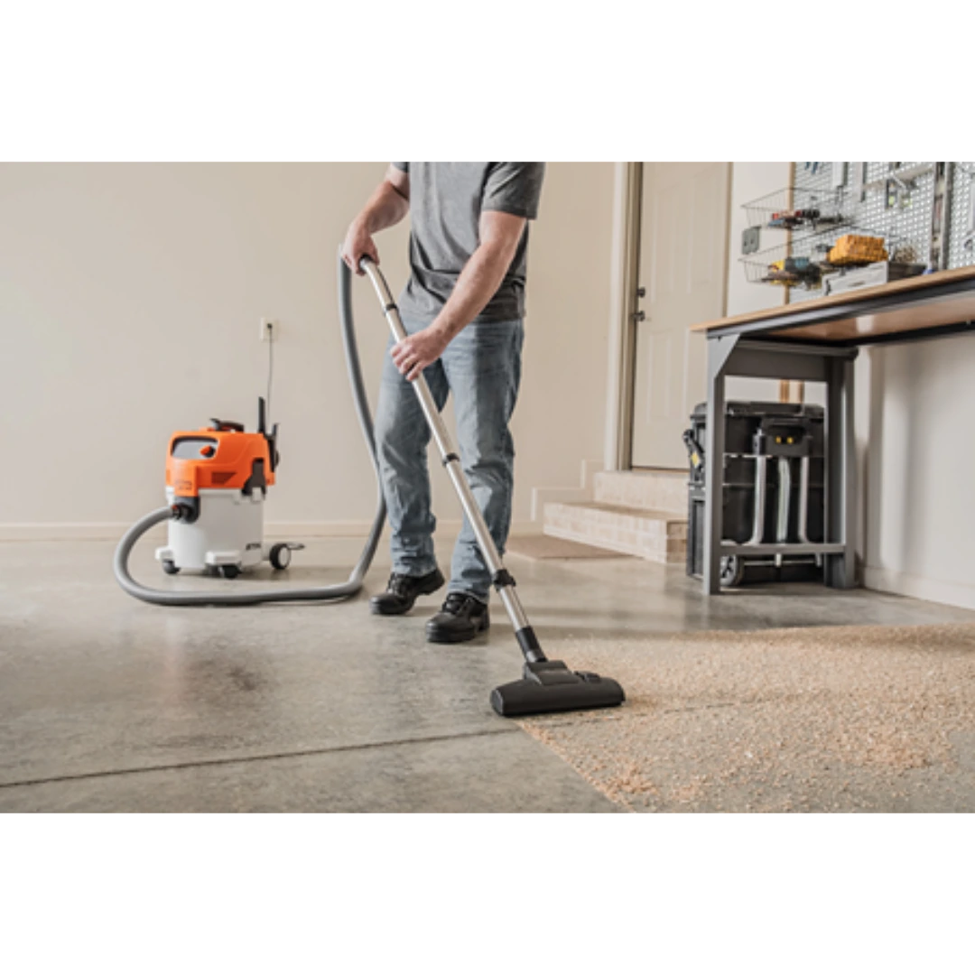 Person using an orange and white wet/dry vacuum with metal extension tube to clean debris on a concrete floor