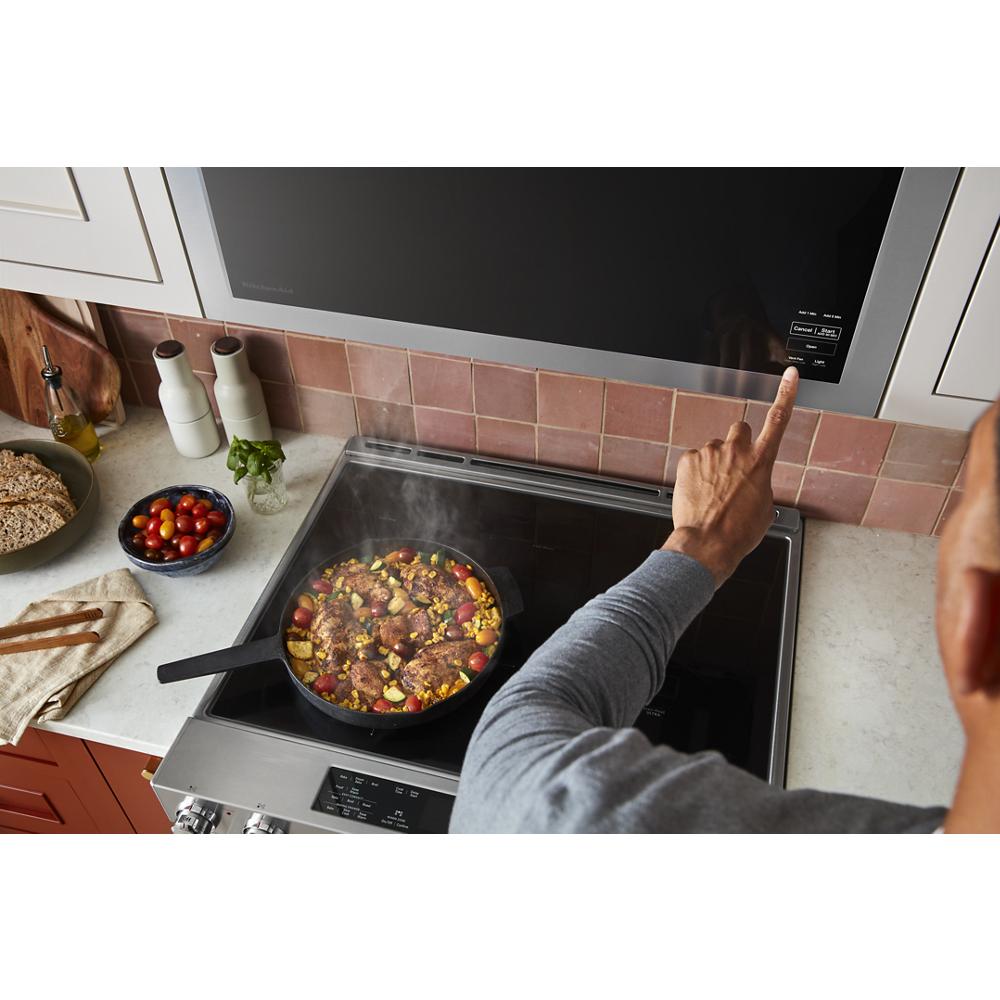 Person using touchscreen control on stainless steel microwave above black glass cooktop with food cooking in skillet