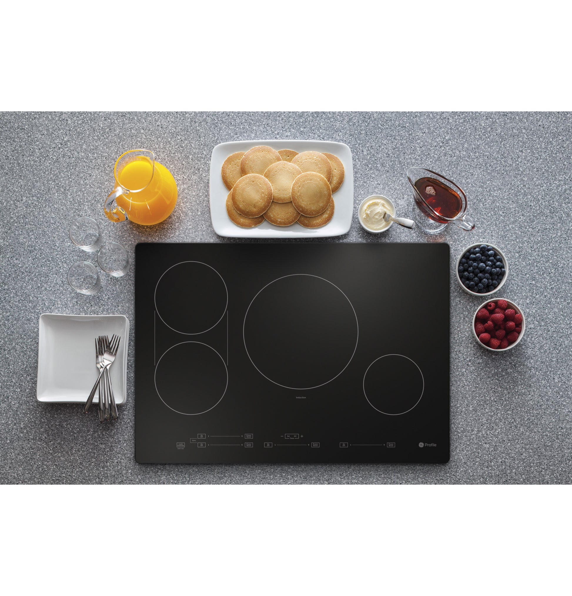 Top-down view of a sleek black induction cooktop with pancakes, syrup, berries, and orange juice on a speckled gray countertop