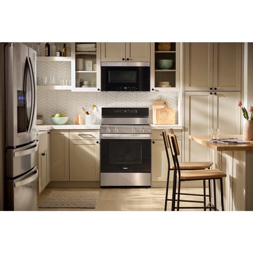 Electric range with digital control and stainless steel finish in modern kitchen with beige cabinetry and wooden dining chairs