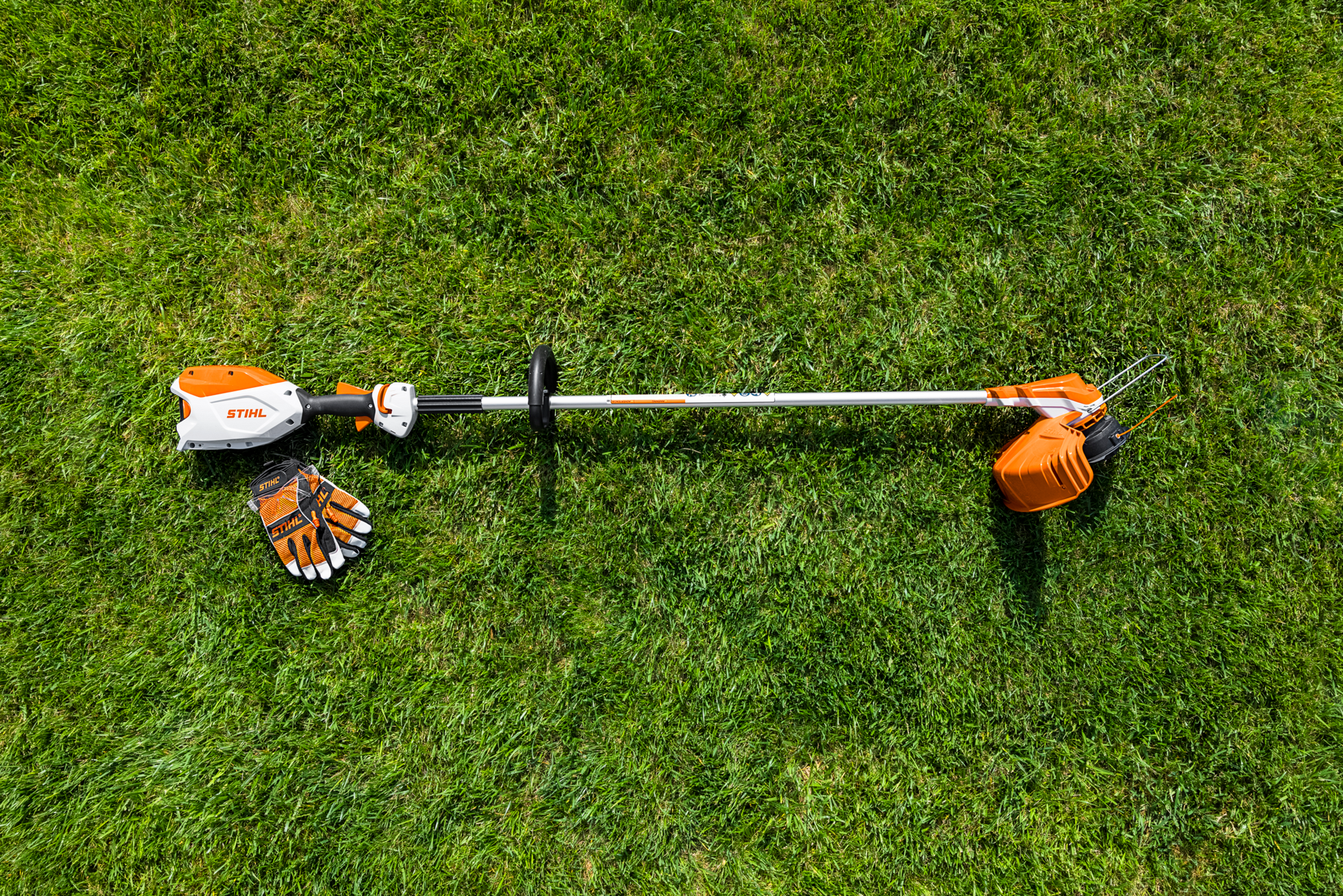 Orange and white battery-powered garden trimmer with protective gloves on green grass for home improvement tasks