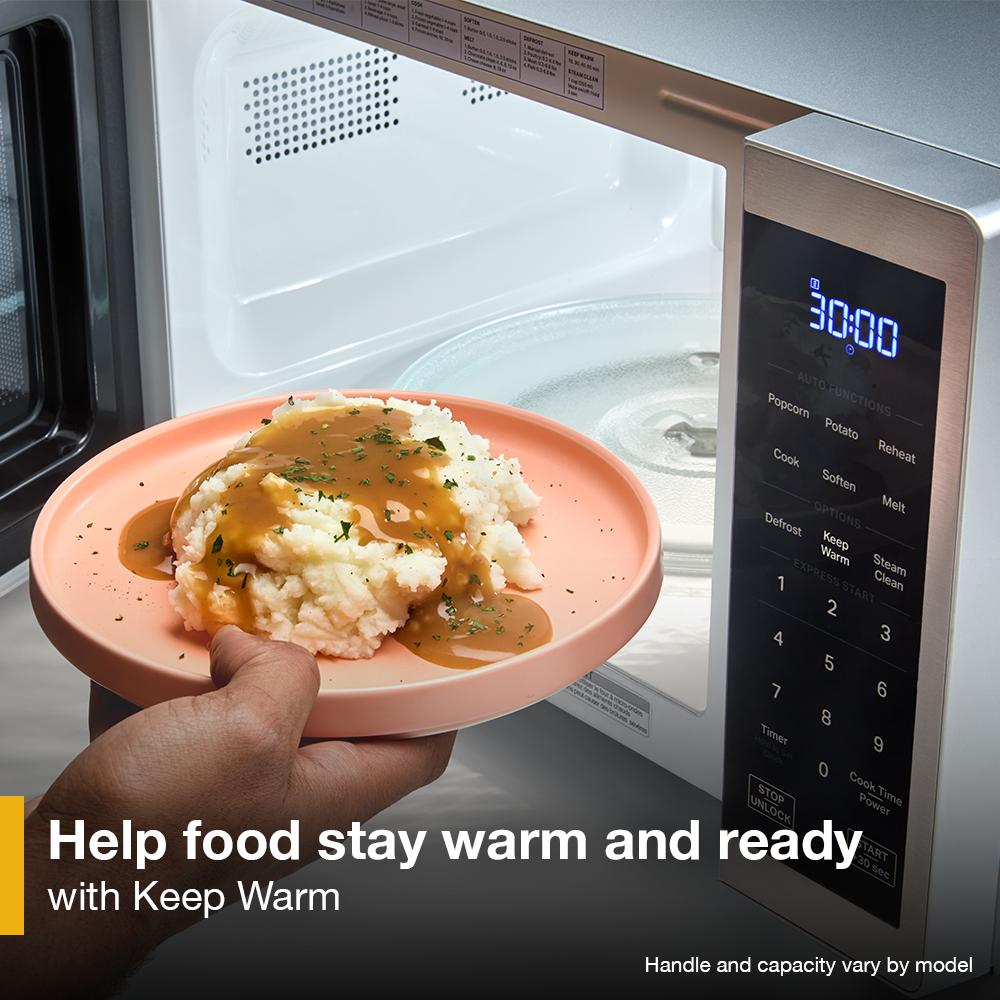 Hand holding plate of mashed potatoes and gravy near open black countertop microwave with digital display and touch controls