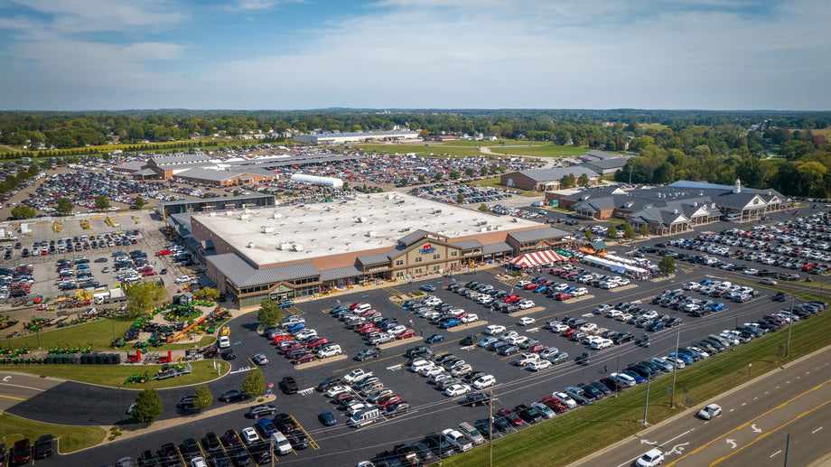Aerial view of a large retail complex with a crowded parking lot, surrounded by trees and additional buildings under a clear sky.