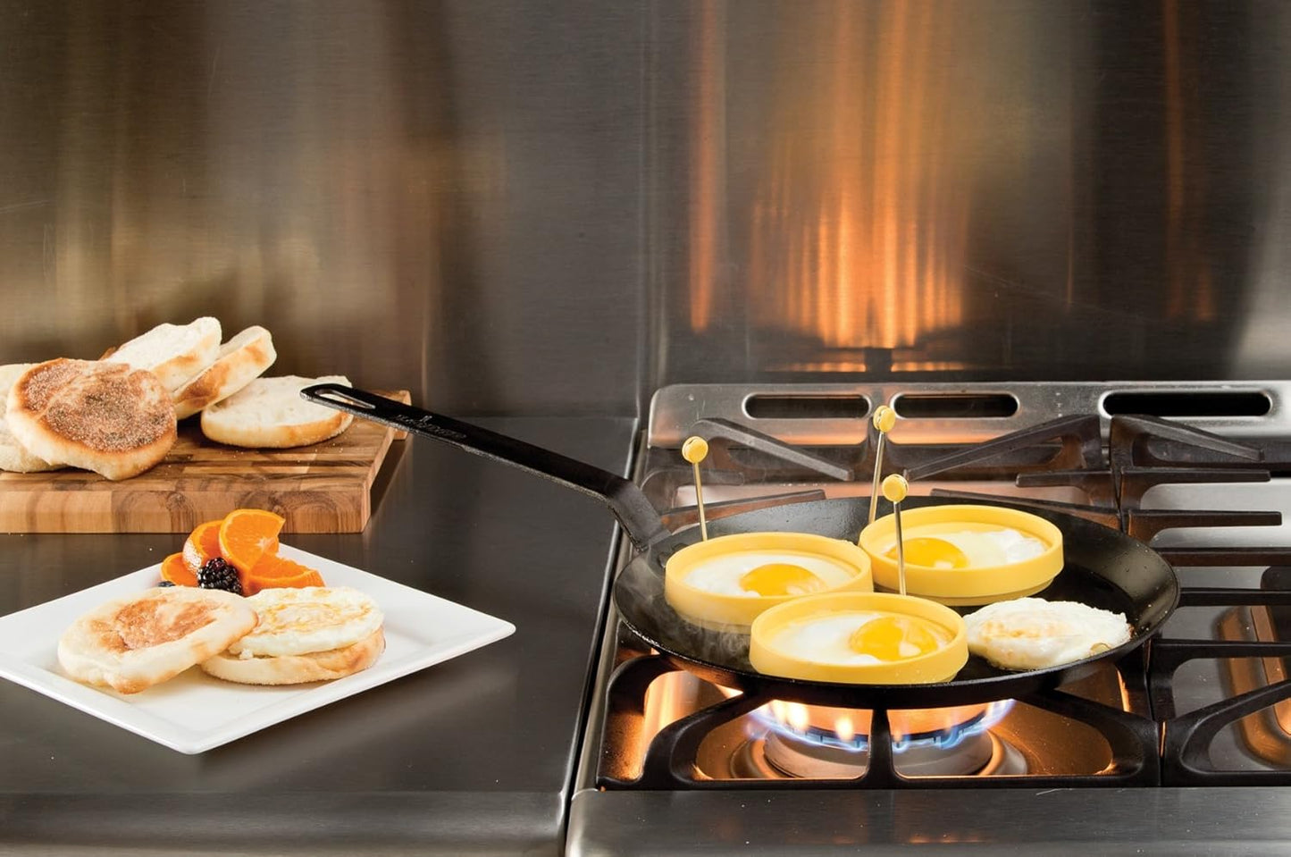 Seasoned steel skillet cooking eggs on gas stove with toasted English muffins on stainless steel countertop