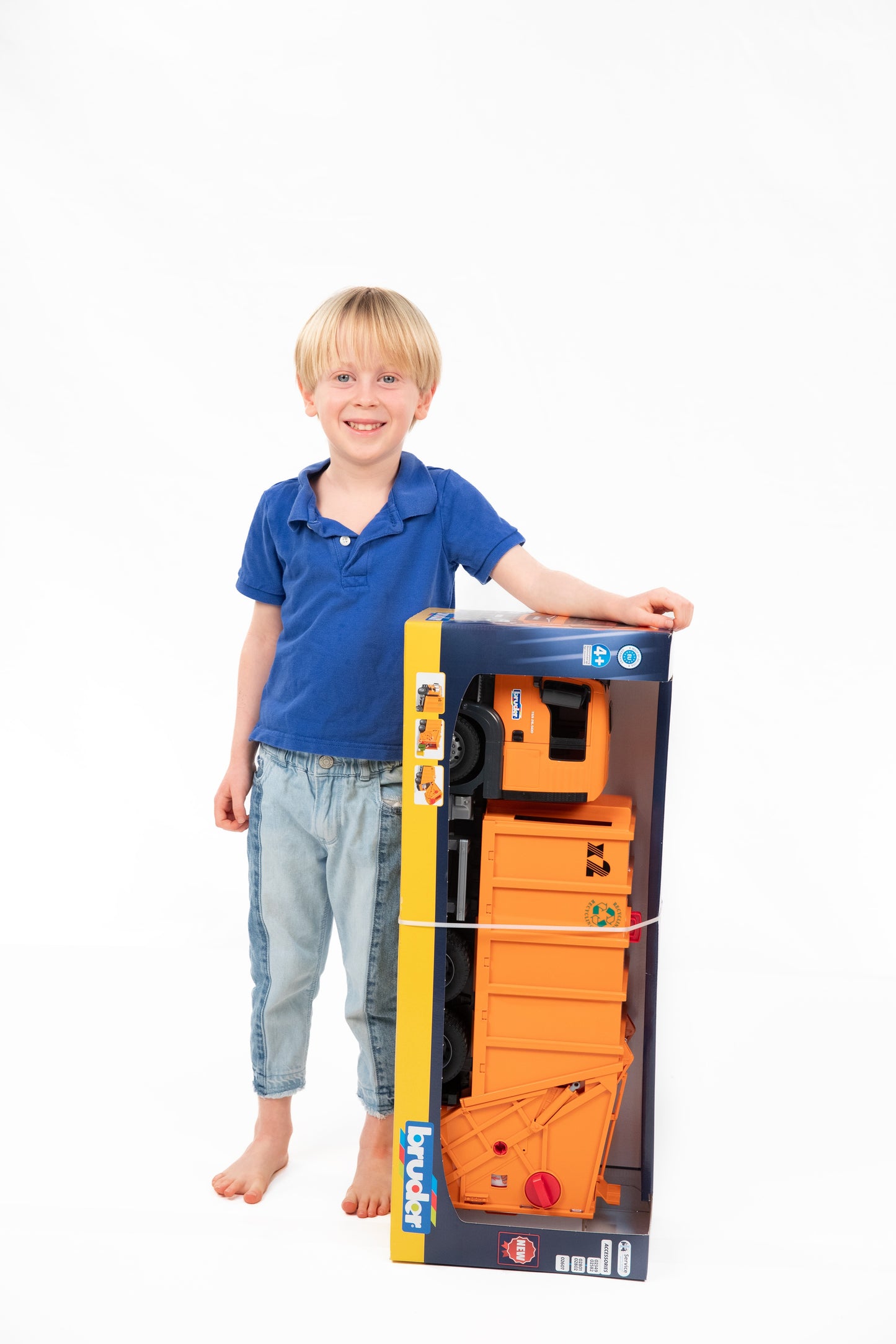 Young boy in blue shirt standing barefoot next to boxed orange Bruder MAN TGS Garbage Truck toy