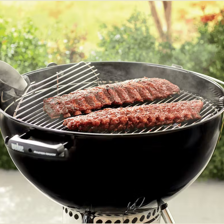 Nickel-plated hinged grill grate on kettle-style charcoal grill with ribs cooking outdoors in garden setting