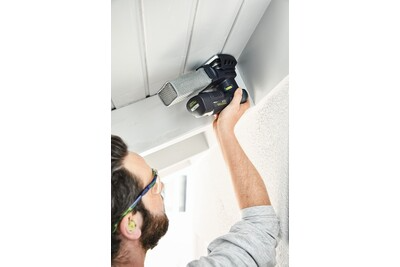 Man wearing safety glasses uses a compact cordless delta sander on a white interior wall corner for home improvement tasks