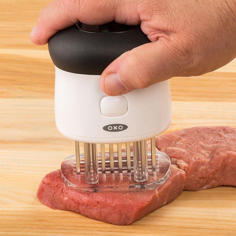 Hand pressing OXO Good Grips bladed tenderizer on steak on wooden surface in kitchen utensil collection under $50